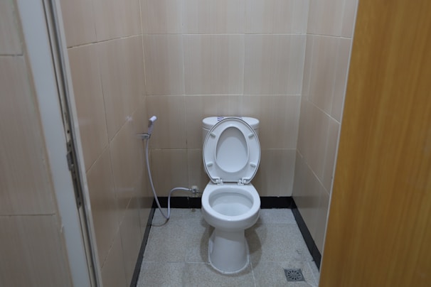 A small bathroom featuring a white toilet with an open lid. Beige tiles cover the walls, and a handheld bidet spray is attached to the wall beside the toilet. The floor is tiled in a light gray color with a small drainage grate near the toilet.