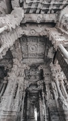 Elaborate stone carvings adorn the pillars and ceiling of an ancient architectural structure, displaying intricate detailing and craftsmanship. The perspective reveals a series of ornately decorated columns leading into the depths of the building, showcasing the cultural and historical significance of the architecture.