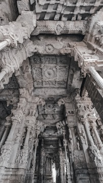 Elaborate stone carvings adorn the pillars and ceiling of an ancient architectural structure, displaying intricate detailing and craftsmanship. The perspective reveals a series of ornately decorated columns leading into the depths of the building, showcasing the cultural and historical significance of the architecture.