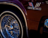 A close-up view of a classic car's glossy maroon body featuring a metallic emblem with a checkered flag and insignia reading '396 Turbo-Jet'. The chrome wire wheel with its intricate design and reflective rim stands out prominently.