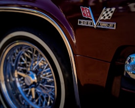 A close-up view of a classic car's glossy maroon body featuring a metallic emblem with a checkered flag and insignia reading '396 Turbo-Jet'. The chrome wire wheel with its intricate design and reflective rim stands out prominently.