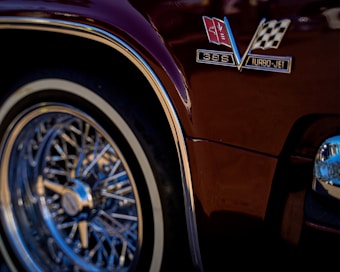 A close-up view of a classic car's glossy maroon body featuring a metallic emblem with a checkered flag and insignia reading '396 Turbo-Jet'. The chrome wire wheel with its intricate design and reflective rim stands out prominently.