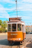 A vintage brown tramcar is stationary on a street track under a bright blue sky. The tramcar has a sign that reads 'Hills Tramcar Tour.' It is adorned with flags on the side, and there are overhead wires connected to its roof. The background features trees and buildings with facades in light colors.