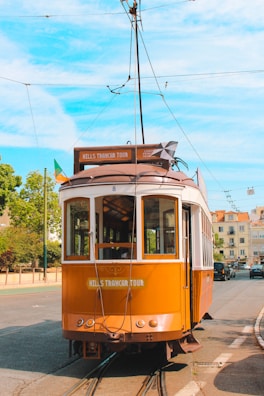 A vintage brown tramcar is stationary on a street track under a bright blue sky. The tramcar has a sign that reads 'Hills Tramcar Tour.' It is adorned with flags on the side, and there are overhead wires connected to its roof. The background features trees and buildings with facades in light colors.