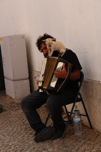 A man with curly hair is sitting on a folding chair, playing an accordion. A small dog is perched on his shoulder, looking forward. The scene takes place against a wall on a cobblestone street. There is a plastic bottle of water on the ground beside him.