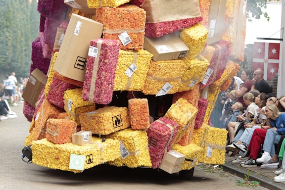 A large, creatively arranged float is covered in blocks made of densely packed colorful flowers, interspersed with cardboard boxes. The float is part of a parade, with people sitting and standing along the street observing the spectacle. The colors of the flowers range from vibrant yellows to deep pinks and reds. The setting appears festive and lively.