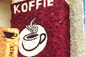 A decorative arrangement of small flowers forms the design of a coffee package labeled with the word 'KOFFIE'. The central focus is a white and dark brown cup of steaming coffee, crafted in contrast with deep red flowers. To the left, there's another coffee-themed design on a yellow-orange background.