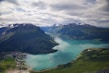 A scenic landscape featuring a serene fjord surrounded by lush green mountains. The water is a calm turquoise-blue, and there are two cruise ships visible—one in the foreground and another in the distance. Small settlements are scattered along the coastline, and patches of snow cap the peaks of the distant mountains under a partly cloudy sky.