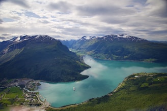 A scenic landscape featuring a serene fjord surrounded by lush green mountains. The water is a calm turquoise-blue, and there are two cruise ships visible—one in the foreground and another in the distance. Small settlements are scattered along the coastline, and patches of snow cap the peaks of the distant mountains under a partly cloudy sky.