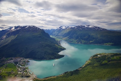 A scenic landscape featuring a serene fjord surrounded by lush green mountains. The water is a calm turquoise-blue, and there are two cruise ships visible—one in the foreground and another in the distance. Small settlements are scattered along the coastline, and patches of snow cap the peaks of the distant mountains under a partly cloudy sky.