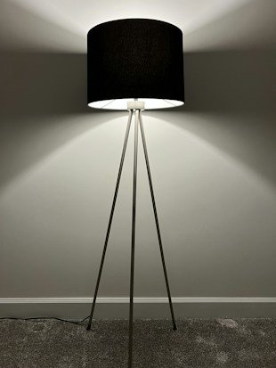 A modern floor lamp with a black cylindrical shade supported by three slender metallic legs. The lamp is positioned against a plain wall, casting soft, radiant light that creates gentle shadows on the wall. The floor is carpeted, and the lamp cord is visible.