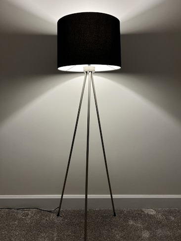 A modern floor lamp with a black cylindrical shade supported by three slender metallic legs. The lamp is positioned against a plain wall, casting soft, radiant light that creates gentle shadows on the wall. The floor is carpeted, and the lamp cord is visible.