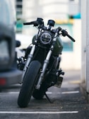 A motorcycle is parked on a street with a slight tilt to the side. The focus is on its front wheel and handlebars, with the background slightly blurred, indicating an urban setting. The motorcycle has a rugged look with a protective grill over the headlight and visible wear on its components.