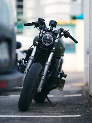 A motorcycle is parked on a street with a slight tilt to the side. The focus is on its front wheel and handlebars, with the background slightly blurred, indicating an urban setting. The motorcycle has a rugged look with a protective grill over the headlight and visible wear on its components.