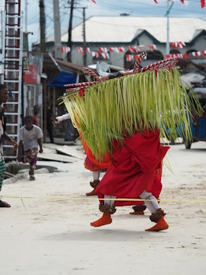 A person wearing a vibrant traditional costume with long green palm leaves covering their upper body and a red skirt. They are participating in a street performance or festival, with a small gathering of people observing. The setting includes local houses with corrugated metal roofs and festive decorations, such as red and white flags.