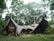 An abstract structure blends into a lush forest environment, constructed from corrugated materials and framed by tall trees. The structure appears to have organic shapes and motifs, including sculptural figures, integrating art with nature.