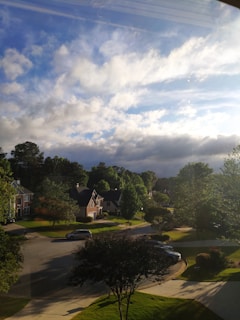 A residential neighborhood under a partly cloudy sky with sunlight streaming through. Several houses with sloping roofs are surrounded by lush green trees. A quiet street curves through the scene, with a few cars parked along the sides. The sky is a mix of blue and dark clouds, suggesting a transition in weather.