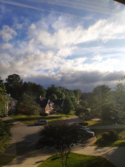 A residential neighborhood under a partly cloudy sky with sunlight streaming through. Several houses with sloping roofs are surrounded by lush green trees. A quiet street curves through the scene, with a few cars parked along the sides. The sky is a mix of blue and dark clouds, suggesting a transition in weather.