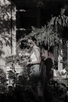 A person wearing sunglasses and a white tank top is holding a potted plant in a shaded garden setting. They are surrounded by various plants and flowers, with sunlight streaming through leaves, casting shadows on the wall and windows behind. The atmosphere is serene and the scene is filled with lush greenery.