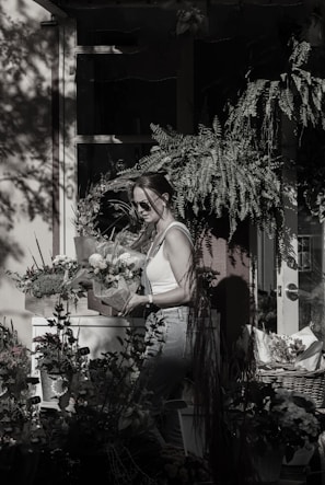 A person wearing sunglasses and a white tank top is holding a potted plant in a shaded garden setting. They are surrounded by various plants and flowers, with sunlight streaming through leaves, casting shadows on the wall and windows behind. The atmosphere is serene and the scene is filled with lush greenery.