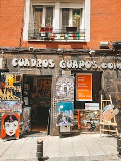 A shop front featuring various artworks displayed outside. The signage reads 'CUADROS GUAPOS.com,' with a variety of colorful illustrations and paintings showcasing cityscapes, stylized portraits, and a large paint palette. The shop is housed in an orange building with potted plants on the balcony.