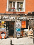 A shop front featuring various artworks displayed outside. The signage reads 'CUADROS GUAPOS.com,' with a variety of colorful illustrations and paintings showcasing cityscapes, stylized portraits, and a large paint palette. The shop is housed in an orange building with potted plants on the balcony.