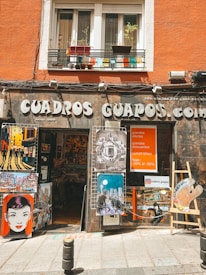 A shop front featuring various artworks displayed outside. The signage reads 'CUADROS GUAPOS.com,' with a variety of colorful illustrations and paintings showcasing cityscapes, stylized portraits, and a large paint palette. The shop is housed in an orange building with potted plants on the balcony.