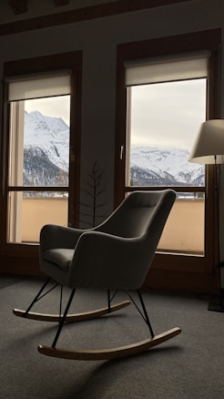 A cozy interior features a rocking chair in a modern design, positioned in front of large windows. The windows reveal a snowy mountain landscape with overcast skies. A floor lamp stands beside the chair, casting soft light into the room.