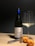 A bottle of wine with a blue foil top stands upright next to a partially filled wine glass. Beside them, a corkscrew and a cork displaying some writing are placed on a light wooden surface. Two apricots lie near the cork, casting soft shadows.