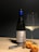 A bottle of wine with a blue foil top stands upright next to a partially filled wine glass. Beside them, a corkscrew and a cork displaying some writing are placed on a light wooden surface. Two apricots lie near the cork, casting soft shadows.