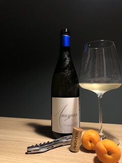 A bottle of wine with a blue foil top stands upright next to a partially filled wine glass. Beside them, a corkscrew and a cork displaying some writing are placed on a light wooden surface. Two apricots lie near the cork, casting soft shadows.