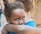 A young child with tears in their eyes clings emotionally to an adult, who is turned away, in an embrace. The background shows a blurred brick structure, suggesting an outdoor setting.
