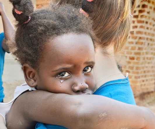 A young child with tears in their eyes clings emotionally to an adult, who is turned away, in an embrace. The background shows a blurred brick structure, suggesting an outdoor setting.