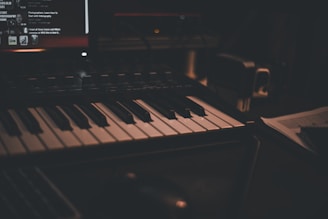 A dimly lit music studio setup featuring a keyboard with black and white keys. In the background, there is a computer screen displaying various controls and text. The environment gives a cozy and intimate ambiance.