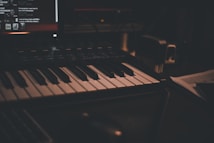 A dimly lit music studio setup featuring a keyboard with black and white keys. In the background, there is a computer screen displaying various controls and text. The environment gives a cozy and intimate ambiance.