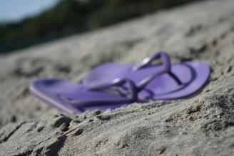 A pair of purple flip-flops lies on sandy ground, with a blurred background of greenery. The image focuses on the flip-flops, highlighting their color and texture against the coarse sand.