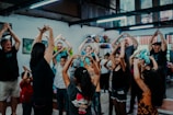 A group of adults and children are gathered indoors in a joyful setting, participating in a physical activity. They are raising their hands above their heads, some holding colorful toys, indicating they might be engaged in an interactive game or exercise. The environment has a relaxed and vibrant atmosphere with bright lighting and creative decorations.