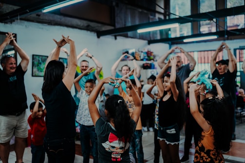 A group of adults and children are gathered indoors in a joyful setting, participating in a physical activity. They are raising their hands above their heads, some holding colorful toys, indicating they might be engaged in an interactive game or exercise. The environment has a relaxed and vibrant atmosphere with bright lighting and creative decorations.