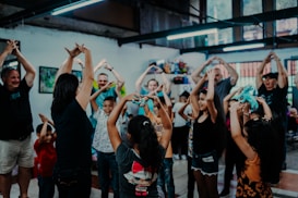 A group of adults and children are gathered indoors in a joyful setting, participating in a physical activity. They are raising their hands above their heads, some holding colorful toys, indicating they might be engaged in an interactive game or exercise. The environment has a relaxed and vibrant atmosphere with bright lighting and creative decorations.