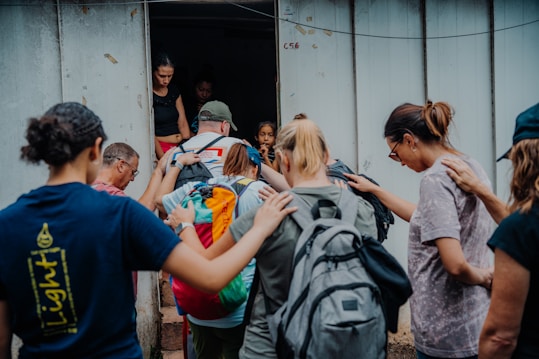 A group of people stands closely together in front of a doorway. Some have their hands on each other's shoulders, showing a sense of support or prayer. They carry backpacks, suggesting they are on a trip or mission.