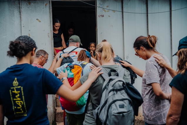 A group of people stands closely together in front of a doorway. Some have their hands on each other's shoulders, showing a sense of support or prayer. They carry backpacks, suggesting they are on a trip or mission.