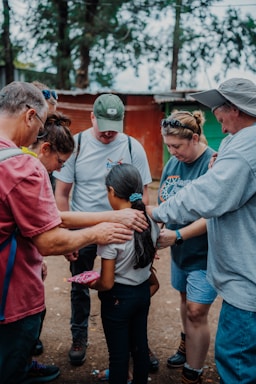 Several adults are gathered around a young girl, placing their hands gently on her shoulders in a supportive gesture. They seem to be outdoors, with trees and a small building visible in the background. The scene conveys a sense of care and community.