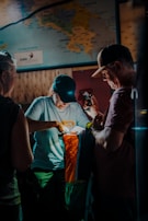 Two people are gathered indoors under dim lighting, examining a colorful outdoor bag labeled 'Cotopaxi'. A third person is partially visible. There's a map on the wall in the background, adding to the exploratory theme.