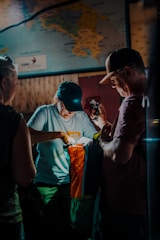 Two people are gathered indoors under dim lighting, examining a colorful outdoor bag labeled 'Cotopaxi'. A third person is partially visible. There's a map on the wall in the background, adding to the exploratory theme.