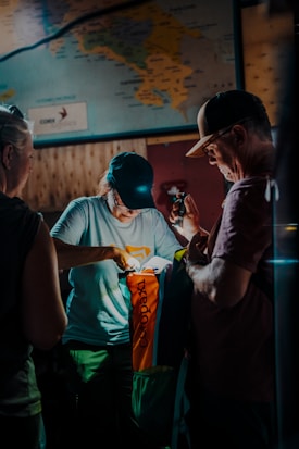 Two people are gathered indoors under dim lighting, examining a colorful outdoor bag labeled 'Cotopaxi'. A third person is partially visible. There's a map on the wall in the background, adding to the exploratory theme.