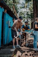 A group of people engaged in a manual task outdoors. One person in the foreground is using a tool, possibly a mattock, while others work in the background near a blue-painted structure. There are trees in the background, suggesting a natural setting.