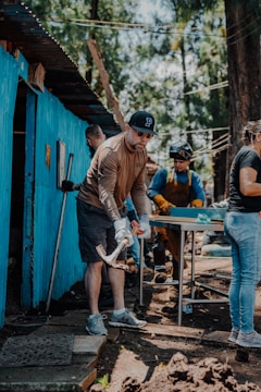 A group of people engaged in a manual task outdoors. One person in the foreground is using a tool, possibly a mattock, while others work in the background near a blue-painted structure. There are trees in the background, suggesting a natural setting.