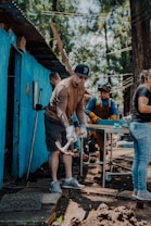 A group of people engaged in a manual task outdoors. One person in the foreground is using a tool, possibly a mattock, while others work in the background near a blue-painted structure. There are trees in the background, suggesting a natural setting.