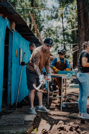 A group of people engaged in a manual task outdoors. One person in the foreground is using a tool, possibly a mattock, while others work in the background near a blue-painted structure. There are trees in the background, suggesting a natural setting.