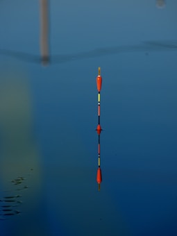 A fishing float stands upright in calm water, perfectly mirrored by its reflection. The water is serene with a deep blue hue, and subtle ripples can be seen around the float. The bright orange and yellow colors of the float contrast with the blue surroundings.